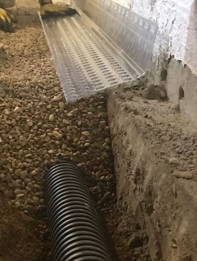 Behncke Construction interior drain tile installation in a Quad Cities basement: bare perforated corrugated pipe with no filter sock, set in a deep bed of clean washed stone along the footing, with a dimpled plastic drainage board running up the foundation wall.
