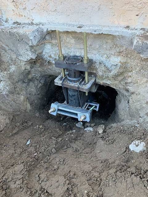 Helical pier bracket and threaded rods installed beneath a foundation footing in a hand-dug pit