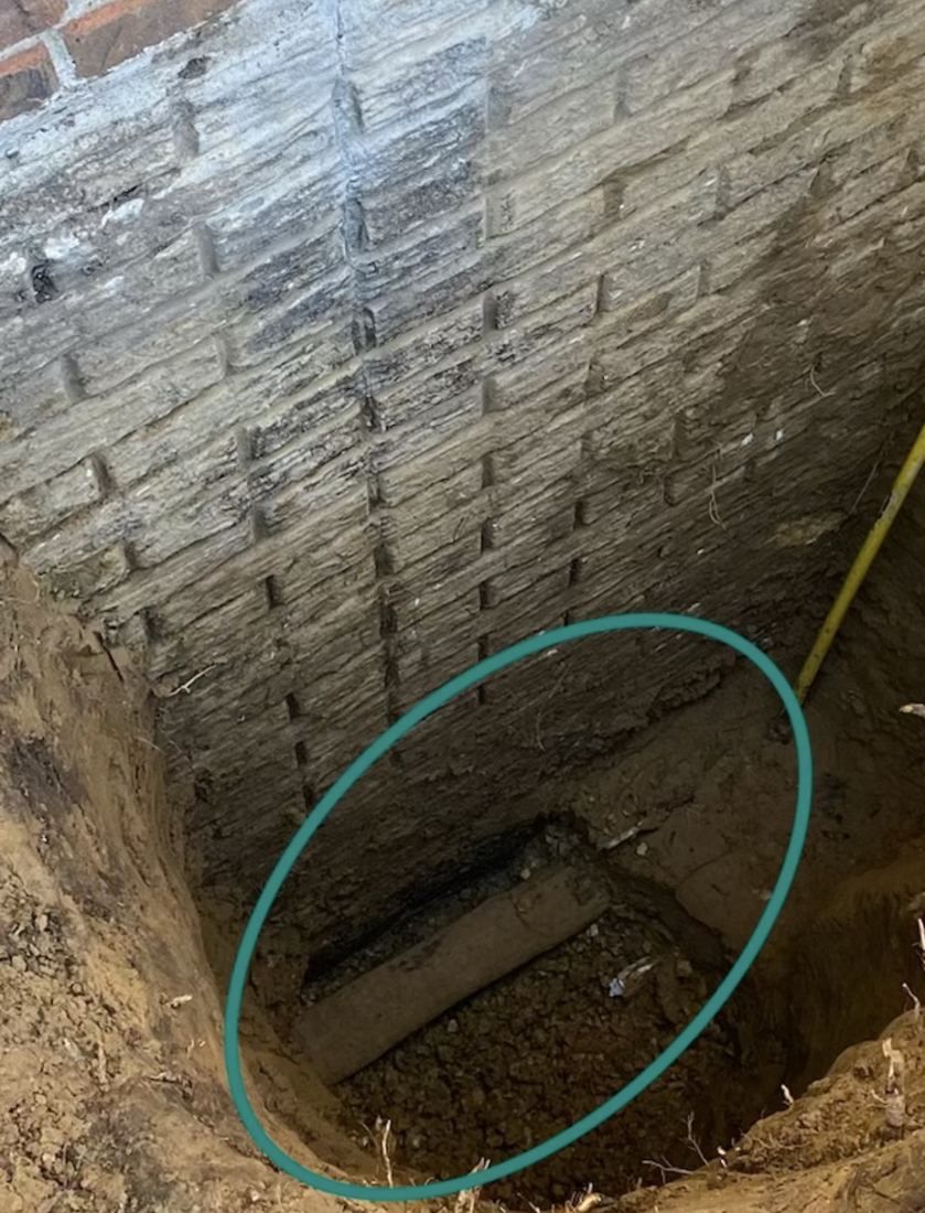 Behncke Construction excavation beside a rock-faced concrete block foundation wall in the Quad Cities, showing the existing exterior footing drain tile circled at the base of the wall, surrounded by packed soil with no visible bed of clean washed stone.