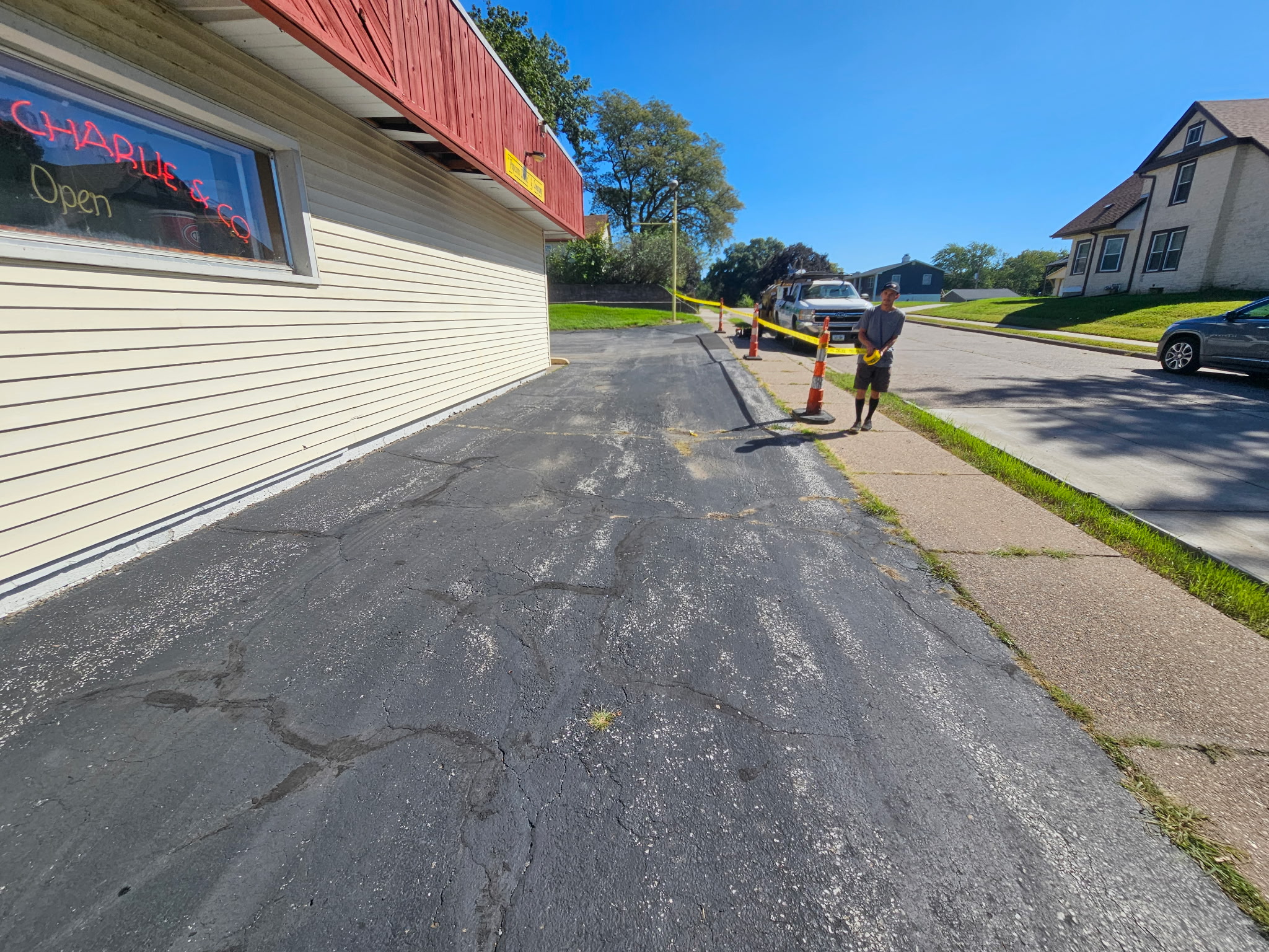 Cracked, weathered asphalt along the side of the Popcorn Charlie's building before sealcoating, with a crew member and truck in the background